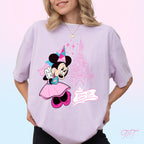 A woman wearing a light lavender oversized t-shirt featuring a Minnie Mouse graphic with a pink castle and 'Dream Big, Dream True' text, set against a pastel pink and blue gradient background.